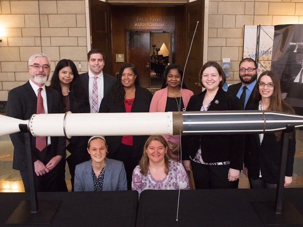 A diverse group of ten people stands and kneels behind a full-scale model of the Explorer 1 satellite
